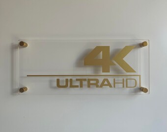 May include: A clear acrylic sign with gold lettering that reads "4K UltraHD". The sign has four gold metal brackets.