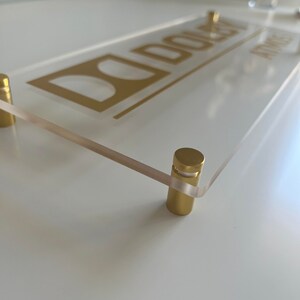 May include: A clear acrylic sign with the name "DOLEY" printed in gold lettering. The sign is mounted with two gold metal standoffs.