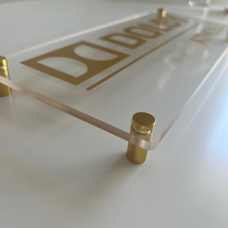 May include: A clear acrylic sign with the name "DOLEY" printed in gold lettering. The sign is mounted with two gold metal standoffs.