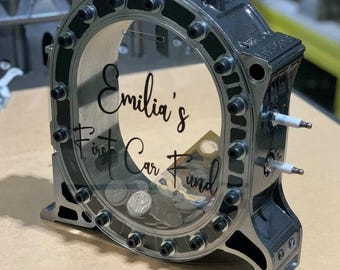 May include: A black metal piggy bank shaped like a car engine with a clear plastic window. The window shows coins inside and a sign that says "Emilia's First Car Fund".