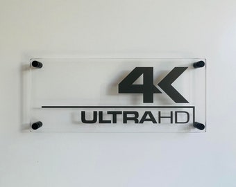 May include: A clear acrylic sign with black lettering that reads "4K ULTRA HD" with black metal mounting brackets.