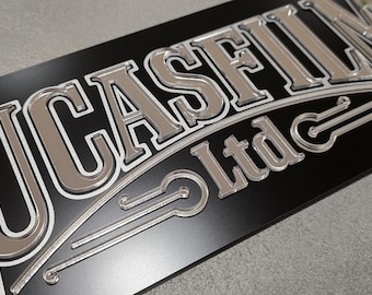 May include: A black rectangular sign with the words "LUCASFILM Ltd" in silver, outlined in white. The letters are raised, creating a 3D effect. The sign has a glossy finish.