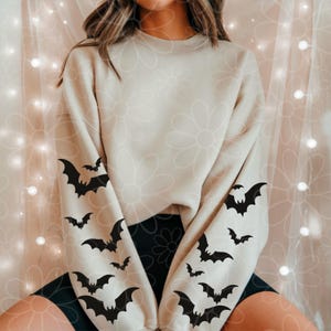 Puede incluir: Una sudadera de cuello redondo en color beige con mangas largas decoradas con un patrón repetitivo de siluetas de murciélagos negros. La prenda cuenta con cuello, puños y dobladillo acanalados, con los gráficos distribuidos en las mangas.