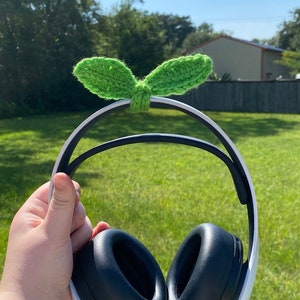 May include: Black and white headphones with a green crocheted sprout attached to the headband. The headphones have black earcups and a white frame. The sprout is made of green yarn and has two leaves.