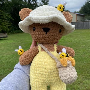 May include: A handmade crochet teddy bear wearing a white hat and yellow overalls. The bear has brown fur, black eyes, and a black nose. It has two small bees attached to the hat and arm, and a honey pot bag.