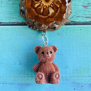 May include: A pendant featuring a polished, dark brown pinecone slice with a symmetrical, flower-like wood grain pattern. A small, textured, light brown teddy bear figurine hangs from the bottom via a silver-toned metal jump ring and bail.