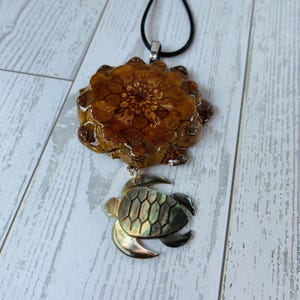 May include: A handcrafted pendant necklace featuring a large, brown, flower-shaped resin pendant with a detailed center. Below, a small, iridescent turtle charm hangs from a silver-toned bail. The necklace has a black cord.