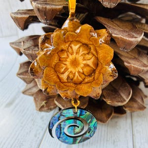 May include: A handcrafted pendant featuring a natural pinecone design with a gold-colored bail and a green cord. The pendant is adorned with a spiral-shaped abalone shell charm, showcasing iridescent blues and greens.