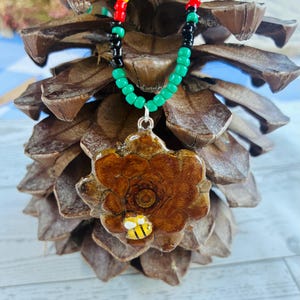 May include: A necklace featuring a brown, polished pinecone slice pendant with a small yellow and black bee charm attached. The pendant hangs from a beaded chain made of small, round, multi-coloured beads in red, yellow, green, and black.