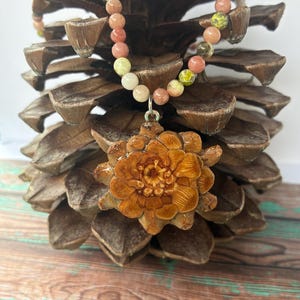 Pode incluir: Um colar com um pingente em forma de flor e uma corrente de contas. O colar tem um pingente em forma de flor e uma corrente de pequenas contas redondas em tons de rosa, amarelo e branco. O pingente é marrom e o colar é exibido em uma pinha.