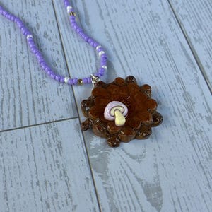 May include: A necklace featuring a string of small purple, white, and gold beads. The pendant is a dark brown, textured, circular piece resembling a pinecone, topped with a small, smooth, light purple mushroom with a cream-coloured stem.