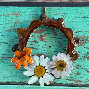 Könnte beinhalten: Ein Anhänger mit einem braunen Harzrahmen, einer orangefarbenen Blume und zwei Gänseblümchen. Der Anhänger hat eine goldfarbene Öse zum Aufhängen. Die Blumen sind in klarem Harz eingeschlossen.