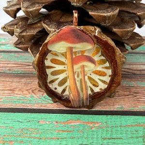 May include: A pendant featuring a mushroom design. The pendant has a copper-colored bail and a resin-coated pinecone base. The mushroom illustration has red caps and tan stems, set against a gold and white patterned background.