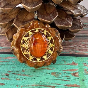 Peut inclure: Pendentif artisanal avec une grosse pierre polie de couleur ambre sertie dans un motif d'éclat d'étoile doré. Le pendentif est enfermé dans un cadre texturé, brun, de forme organique, suspendu à une bélière argentée.