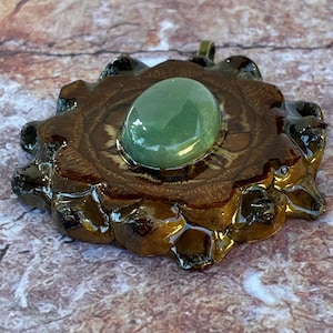 Puede incluir: Colgante artesanal con una gran piedra preciosa verde ovalada engastada en una base de resina marrón en forma de flor. La resina tiene un acabado brillante y un diseño tallado detallado. Se adjunta una pequeña argolla dorada para colgar.