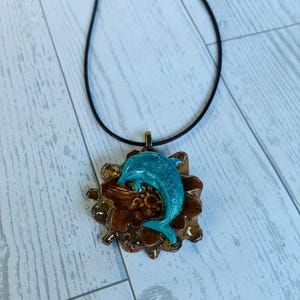 May include: A necklace featuring a black cord and a pendant composed of a brown, textured pinecone-like base topped with a glossy, bright turquoise dolphin figure. The dolphin is positioned in a curved, leaping motion over the brown base.