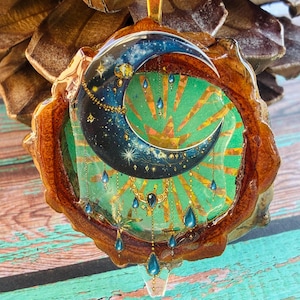 May include: A circular pendant featuring a dark blue crescent moon with gold star accents and dangling blue teardrop charms set against a bright green background with radiating gold sunburst lines, all encased in a polished natural wood frame.