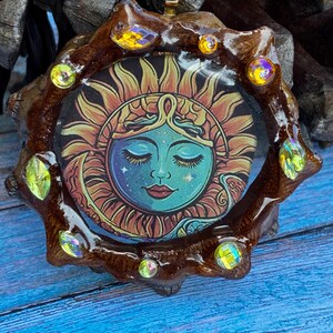 Puede incluir: Colgante artesanal con un diseño de sol con una cara serena en el centro. El sol está rodeado por un marco marrón texturizado adornado con cristales iridiscentes. El colgante tiene una fianza dorada.
