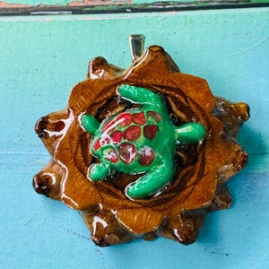 Peut inclure: Un pendentif composé d'une base en écaille de pomme de pin brune, texturée et brillante, avec une figurine de tortue de mer verte en relief au centre. La carapace de la tortue présente des taches circulaires rouges. Une bélière argentée est fixée.