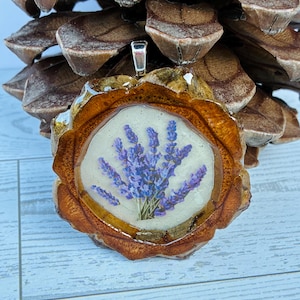 Puede incluir: Colgante artesanal con un ramo de lavanda encapsulado en resina. El colgante está engarzado en un marco de madera natural con una fianza plateada. En el fondo, una piña.