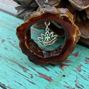 Pode incluir: Pingente de madeira artesanal com uma moldura circular marrom escuro. O pingente apresenta um charme de flor de lótus dourada com uma figura sentada. O fundo é uma superfície turquesa desgastada.