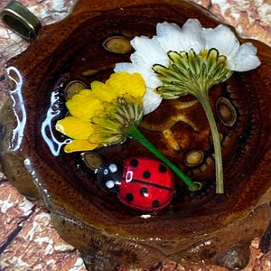 Könnte beinhalten: Handgefertigter Anhänger mit einem Marienkäfer, gelben und weißen Blumen und Samen, die in klarem Harz eingeschlossen sind. Der Anhänger ist in einer dunkelbraunen, holzähnlichen Basis mit einer silberfarbenen Öse zum Aufhängen gefasst. Der Marienkäfer ist rot mit schwarzen Punkten.