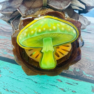 May include: A pendant featuring a bright green mushroom illustration encased in a clear resin layer, set within a dark brown natural pinecone scale frame. The mushroom has a wide cap with white spots and a central stem, topped with a metal bail.