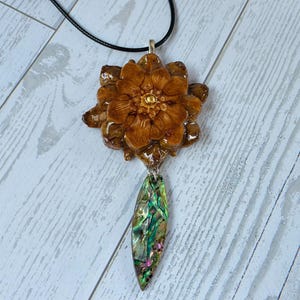 May include: A handcrafted pendant necklace featuring a detailed, brown flower pendant with a gold center. Below the flower hangs an iridescent, oval-shaped abalone shell. The necklace has a black cord.