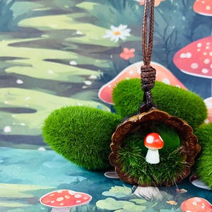 以下が含まれることがあります： 緑色の人工苔で満たされたダークブラウンの質感のある円形の木製フレームの中に、赤と白のミニチュアキノコのフィギュアが収められたペンダントネックレス。ダークブラウンの編み込みコードで吊り下げられています。