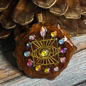 May include: A handcrafted pendant featuring a golden eye design with colorful beaded accents. The pendant is set on a piece of wood and resin, with a gold-colored bail. The background is a pinecone.
