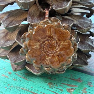 May include: A brown pendant necklace featuring a pine cone design. The pendant is made of a resin-like material with a detailed, textured surface. The necklace has a brown cord and a copper-colored clasp. The pendant is approximately 2 inches in diameter.