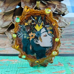 May include: A circular pendant featuring a profile illustration of a person with a floral crown, encased in a brown textured frame adorned with small, round, iridescent and amber-colored crystals, topped with a bronze-toned metal bail.