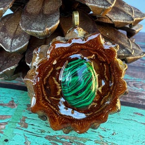 以下が含まれることがあります： 樹脂でコーティングされた松ぼっくりのベースに、緑と黒のマラカイトの石をセットした手作りのペンダント。ペンダントには金色のベイルが付いています。松ぼっくりは濃い茶色です。