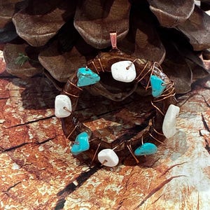 May include: A handcrafted pendant featuring a circular design. The pendant is made with copper wire and adorned with turquoise and white stones. The pendant is displayed against a backdrop of a pine cone.