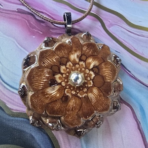 May include: A pendant necklace featuring a detailed, brown, flower-shaped pendant with a silver-colored bail. The pendant has a central, clear, round gem and is suspended from a thin, tan cord. The pendant's design resembles a pinecone.