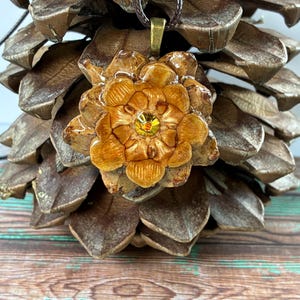 May include: A handcrafted pendant necklace featuring a detailed, flower-shaped pendant with a central yellow gemstone. The pendant is a warm brown color, suspended from a brown cord. The background includes a pinecone and a wooden surface.