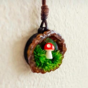 Könnte beinhalten: Ein Anhänger bestehend aus einer braunen Walnussschalenhälfte mit einem Miniatur-Pilz in Rot und Weiß. Der Pilz ist von leuchtend grünem Kunstmoos umgeben. Der Anhänger hängt an einer dunkelbraunen Kordel mit einem verknoteten Verschluss.