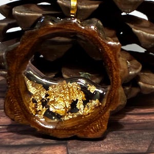 May include: A handcrafted pendant with a unique design. The pendant features a wooden frame with a clear resin interior filled with gold leaf flakes. The pendant is suspended from a gold-colored bail.