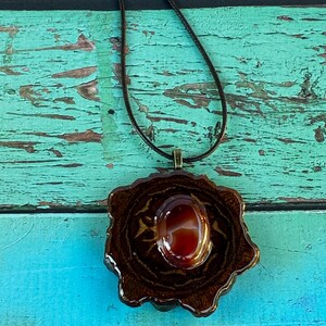 Peut inclure: Un collier pendentif artisanal avec une pierre précieuse brun-rougeâtre ovale, sertie dans une base marron foncé de forme organique. Le pendentif est suspendu à un fin cordon marron foncé. Le collier est présenté sur un fond en bois turquoise et marron.