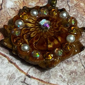 Peut inclure: Un pendentif artisanal avec un motif floral. Le pendentif est marron avec une finition brillante, orné de fausses perles, de cristaux irisés et d'un cristal central multicolore. Il possède une bélière dorée pour la suspension.