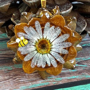 May include: A handcrafted pendant featuring a daisy with white petals and a yellow center, accented by a small bee. The pendant is set on a brown, pinecone-shaped base with a gold-colored bail. The background is a wooden surface.
