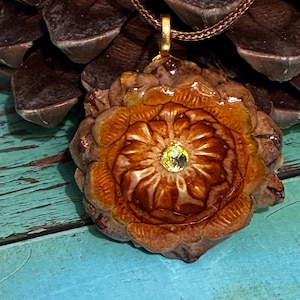 Peut inclure: Un collier pendentif artisanal avec un motif de pomme de pin marron détaillé et une pierre précieuse jaune-vert au centre. Le pendentif est suspendu à une chaîne dorée, mettant en valeur une esthétique naturelle et terreuse. Le collier est un bijou unique.