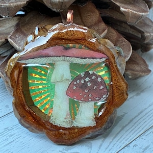 May include: A handcrafted pendant featuring a mushroom design. The pendant has a brown wooden frame with a clear resin coating. The mushroom illustration has a red and white cap with a white stem, set against a green and gold background.