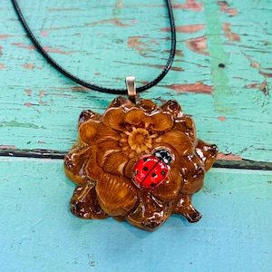 May include: A brown, glossy, flower-shaped pendant made from a pine cone segment, featuring a small, bright red ladybug with black spots attached to the surface. The pendant hangs from a thin, black cord necklace, creating a nature-inspired accessory.