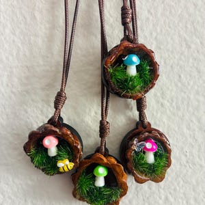 May include: Four circular pendants hanging on brown cords, each featuring a miniature scene inside a dark wood-toned frame. The scenes contain green artificial moss and small colourful mushrooms in blue, pink, and green, with a tiny bee and butterfly.