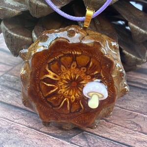 May include: A handcrafted pendant featuring a detailed, brown, pinecone-shaped design with a small, white and yellow mushroom accent. The pendant is suspended from a lavender cord with a gold-colored clasp. The pendant measures approximately 2 inches in diameter.