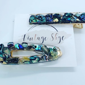 May include: Two hair clips with iridescent abalone shell pieces set in clear resin. One is a rectangular barrette, and the other is an oval clip. The clips have gold-colored metal hardware. The card behind the clips says "Vintage Skye".