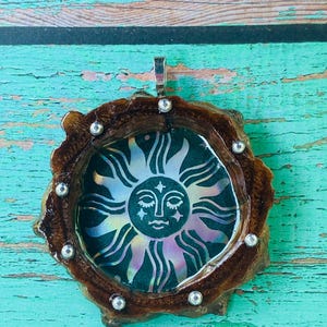 May include: A handcrafted pendant featuring a sun design. The sun has a face and is surrounded by rays of light. The pendant is set in a dark brown, organic-shaped frame with silver accents and a silver bail for hanging.