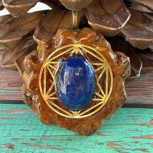 Puede incluir: Un colgante con una gema azul oscuro pulida engastada en un diseño geométrico dorado. El colgante está encerrado en una piña marrón recubierta de resina, creando una pieza natural y artística.