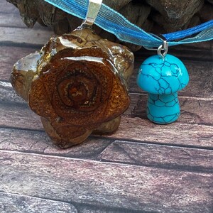 Puede incluir: Dos collares con colgantes. Uno presenta una rodaja de piña marrón recubierta de resina, el otro un hongo turquesa. Ambos cuelgan de una cinta azul. El fondo es una superficie de madera.